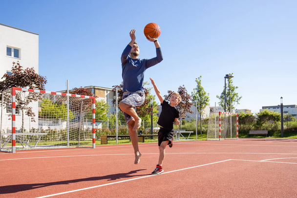 Ο μπαμπάς και ο γιος παίζουν μπάσκετ ξυπόλητοι με την μπάλα σε μια παιδική χαρά - Φωτογραφία, εικόνα