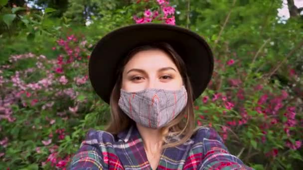 Naisella on suojaava naamio ulkona sepelvaltimoviruksen covid-19 pandemian aikana. Tyttö nauttii haju kukkia kukkiva puutarha kuvaamisen itse. - Materiaali, video