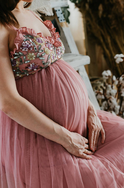 Pregnant woman in pink dress holds hands on belly. Pregnancy, maternity, preparation and expectation concept. Close-up, copy space, intereior. Beautiful tender mood photo of pregnancy. - Photo, image