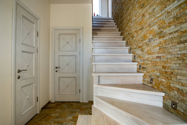 Stylowe drewniane współczesne schody wewnątrz wnętrza domu poddasza. Nowoczesny korytarz z dekoracyjnymi ścianami z cegły wapiennej i schodami z białego dębu. - Zdjęcie, obraz