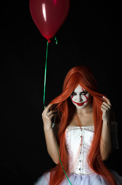 uma menina em um traje de palhaço com maquiagem assustadora e uma bola vermelha em um fundo preto
 - Foto, Imagem