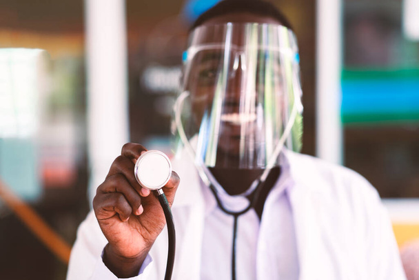 Afrikanischer Arzt trägt Gesichtsschutz und hält Stethoskop freundlich lächelnd in der Hand - Foto, Bild