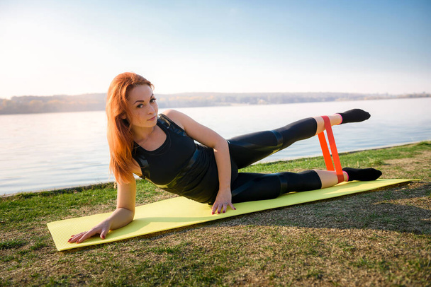  Mujer joven haciendo ejercicios con banda de resistencia al aire libre cerca del lago durante el día. Estilo de vida saludable - Foto, imagen
