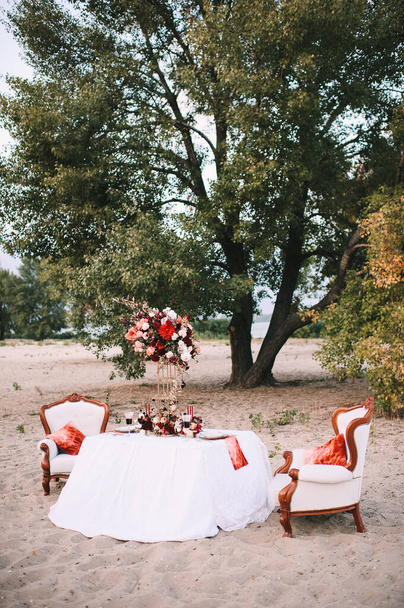 Un banchetto sulla natura, un tavolo con una tovaglia bianca, piatti d'epoca con orlo d'oro, bicchieri di vino, posate, candele, una composizione di fiori, amarilli, rose, dalie rosse
. - Foto, immagini