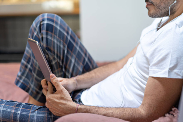 hombre sosteniendo el uso de tableta digital y auriculares en la cama en casa con pijama. Educación online, trabajo, videollamada
 - Foto, imagen