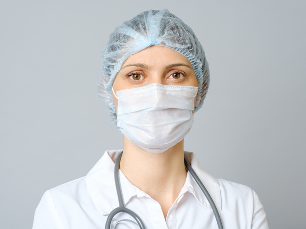 Portrait of young female doctor in protective medical mask on her face and cap on her head. Isolated on gray background - Фото, изображение
