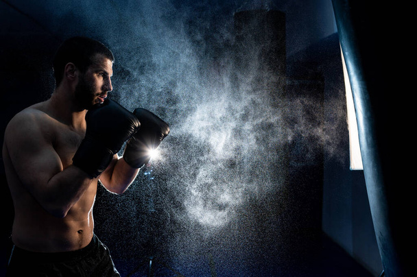  El boxeador tailandés sobre un fondo oscuro golpea una bolsa de boxeo. Publicidad Artes Marciales Fitness Center - Foto, imagen