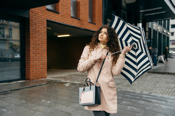 Charmante jeune femme bouclée mestizo sous parapluie marchant dans la rue de la ville de mégapole dans les jours de pluie - Photo, image