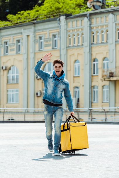 Positive courier with thermo bag waving hand on urban street  - 写真・画像