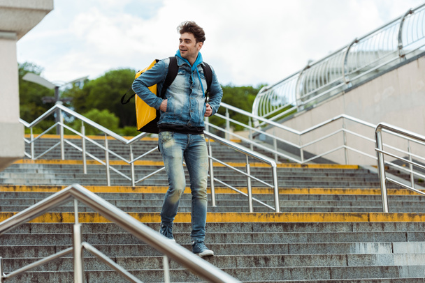 Sırt çantalı gülümseyen kuryenin seçmeli odak noktası merdivenlerde tırabzanla yürüyor.  - Fotoğraf, Görsel
