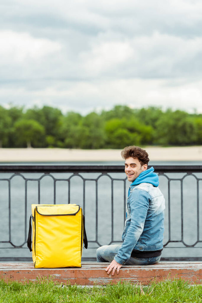 Vista trasera del mensajero sonriente sentado cerca de la bolsa térmica en el banco en el paseo marítimo
 - Foto, Imagen