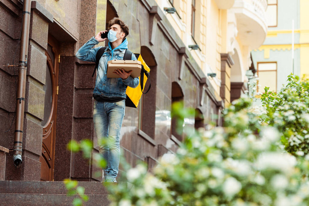 Szelektív fókusz futár orvosi maszk beszél okostelefon és gazdaság pizza dobozok a városi utcán  - Fotó, kép