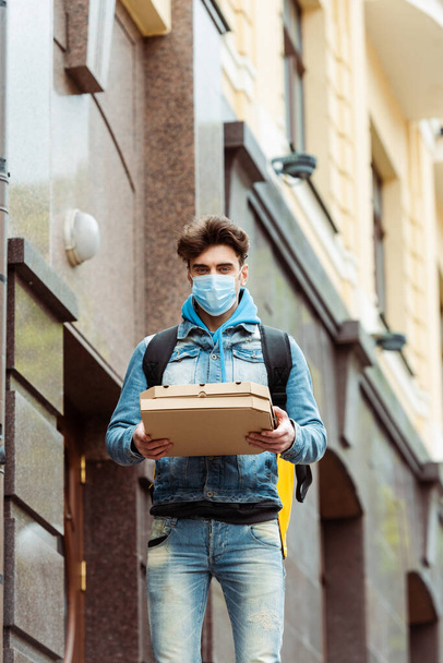 Mensajero en máscara médica mirando a la cámara mientras sostiene cajas de pizza en la calle urbana
 - Foto, imagen