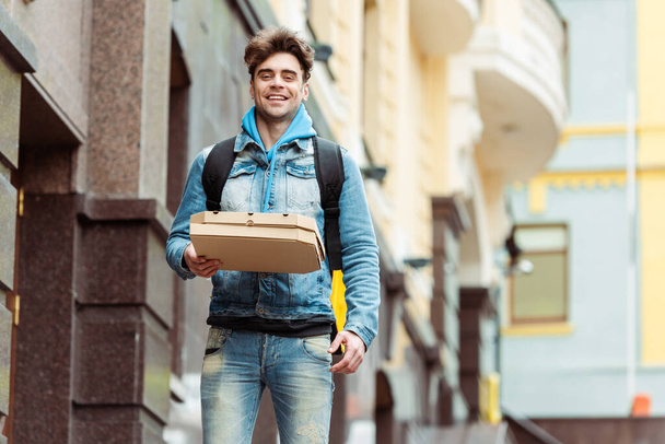 Positive courier with thermo backpack smiling at camera while holding pizza boxes on urban street  - 写真・画像