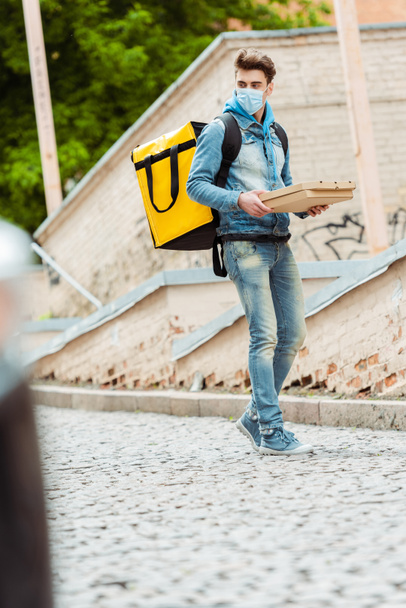 Selective focus of delivery man in medical mask with thermo backpack holding pizza boxes on urban street  - Zdjęcie, obraz