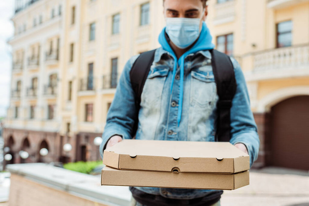 Focus selettivo dell'uomo di consegna in maschera medica che tiene scatole di pizza con strada urbana sullo sfondo
  - Foto, immagini