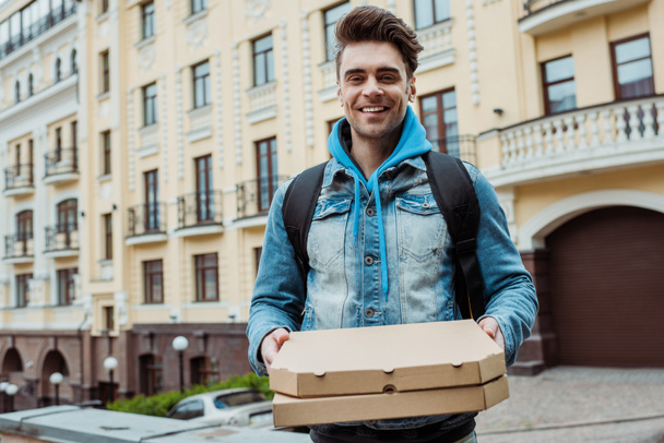 Jóképű futár pizzás dobozokkal a kezében és mosolyog a kamerába a városi utcán.  - Fotó, kép