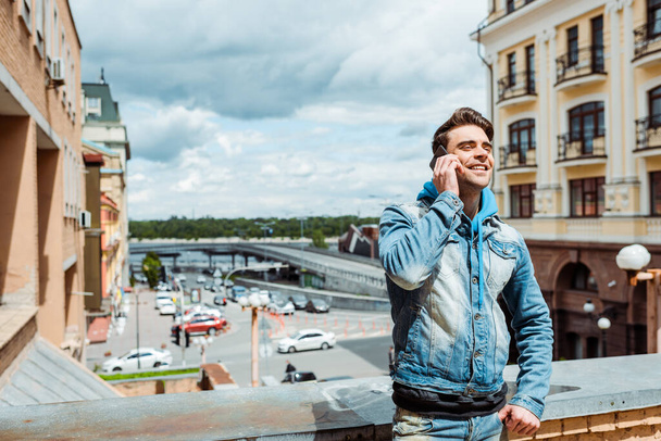 Красивий чоловік посміхається під час розмови на смартфоні з хмарним небом та міською вулицею на фоні
  - Фото, зображення
