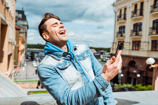 Bonito homem rindo enquanto segura smartphone na rua urbana
  - Foto, Imagem