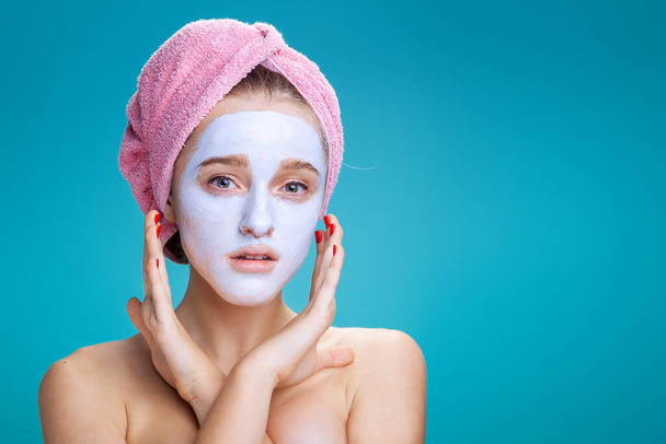 teenager dívka s kosmetickou maskou na tváři a růžový ručník na hlavě na modrém pozadí ve studiu - Fotografie, Obrázek