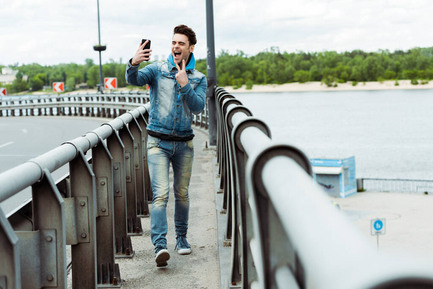 Foco seletivo de homem alegre mostrando sinal de paz ao tomar selfie com smartphone na ponte
  - Foto, Imagem