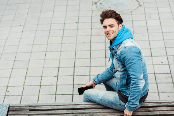 Side view of positive man in earphones smiling at camera while holding smartphone on bench  - Foto, Bild