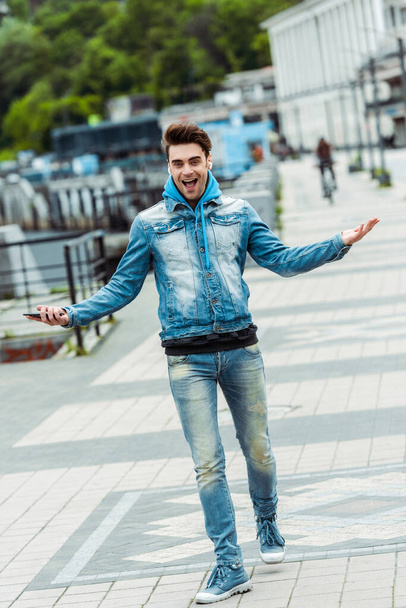 Cheerful man in headphones holding smartphone and smiling at camera on urban street  - Fotoğraf, Görsel