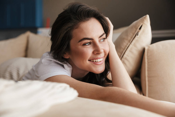 Beautiful smiling young brunette woman relaxing on a couch at home, looking away - Photo, Image
