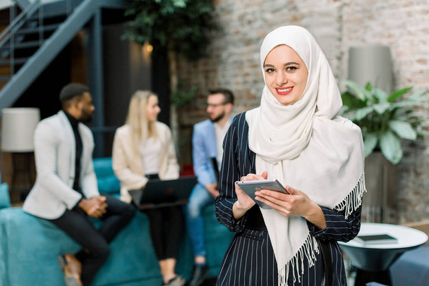 Close up portrait face of attractive muslim woman in white hijab, holding digital tablet, looking at camera and smiling, standing on her multiethnic office colleagues background - Фото, изображение