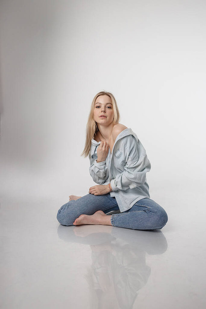 portrait of sexy caucasian woman with long hair posing in blue shirt and jeans on white studio background. model tests of pretty girl in basic clothes. attractive female sitting on floor on her knees - Valokuva, kuva