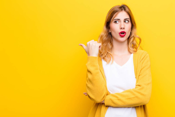young pretty blonde woman looking astonished in disbelief, pointing at object on the side and saying wow, unbelievable against yellow wall - Foto, Bild