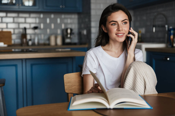 Attractive young brunette woman talking on mobile phone while reading a book while sitting on a kitchen - Фото, зображення