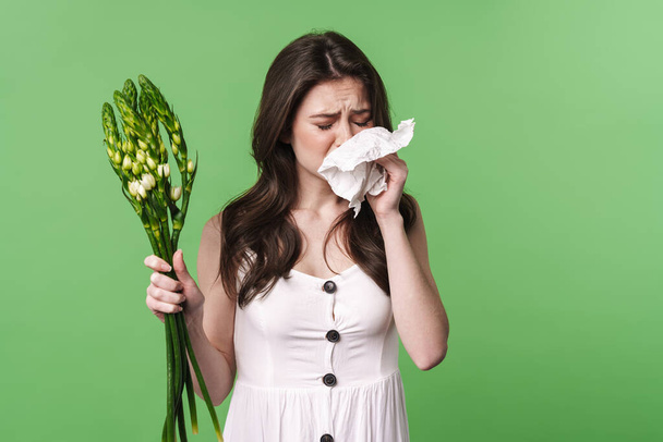 Image of unhappy young woman with allergy holding handkerchief and flowers isolated over green background - Foto, Imagem