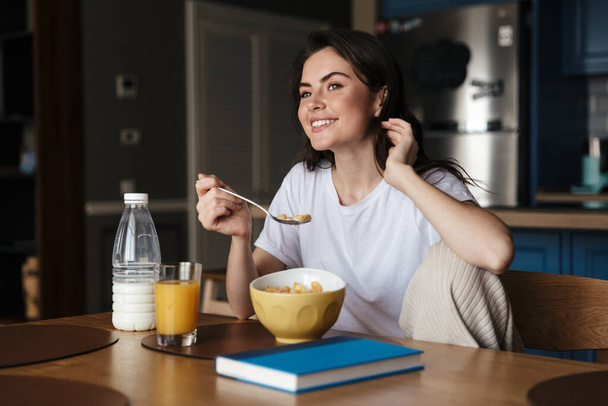 Attractive smiling young brunette woman having a healthy breakfast while sitting at the kitchen table - Valokuva, kuva