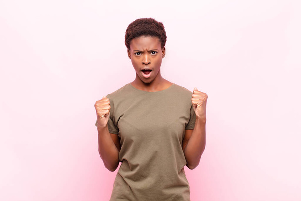 young pretty black womanshouting aggressively with an angry expression or with fists clenched celebrating success against pink wall - Foto, Bild