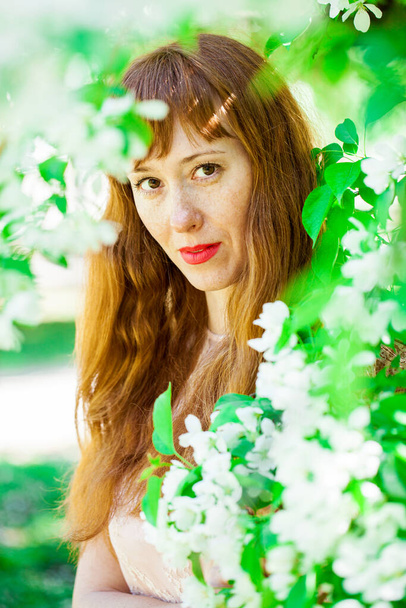 Portrait of a young red-haired woman posing against the backdrop of a blossoming apple tree - Fotografie, Obrázek