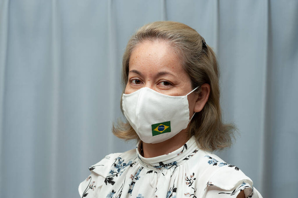 COVID-19 Pandemie - Portrait einer Frau mittleren Alters (Brasilianerin / Japanerin), die eine selbstgemachte chirurgische Gesichtsmaske mit Brasilien-Fahne trägt. Coronavirus-Schutz. Drinnen. Frontansicht.  - Foto, Bild