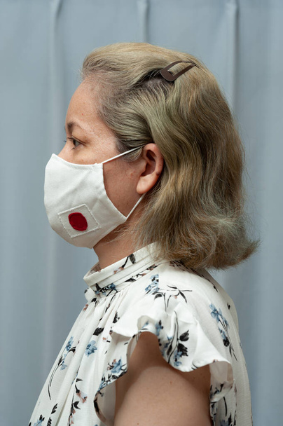 COVID-19 Salgını - Orta yaşlı (Brezilyalı / Japon) kadının DIY ev yapımı cerrahi yüz maskesi ve Japon bayrağı taktığı yan görünüm portresi. Coronavirus koruması. İçeride. Dikey çekim. - Fotoğraf, Görsel