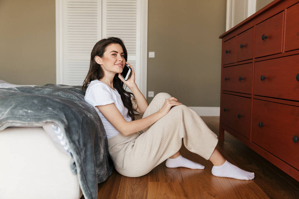Attractive smiling young brunette woman talking on mobile phone while sitting on a floor, leaning on bed at the bedroom - Photo, Image