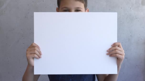 Chłopak trzyma w rękach pusty, biały plakat z makietą. Dzieciak zakrywający twarz deską płócienną. Szara ściana na tle - Materiał filmowy, wideo