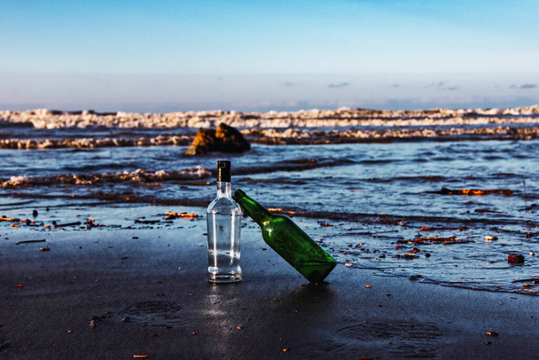 Dwie szklane butelki na brzegu plaży z falami zbliżającymi się do nich. W odniesieniu do środowiska, ponownego wykorzystania, recyklingu, ekologii i opieki nad morzami - Zdjęcie, obraz