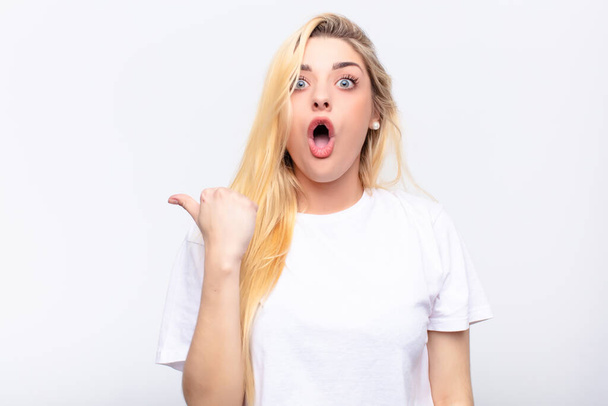 young pretty blonde woman looking astonished in disbelief, pointing at object on the side and saying wow, unbelievable against white wall - Foto, imagen