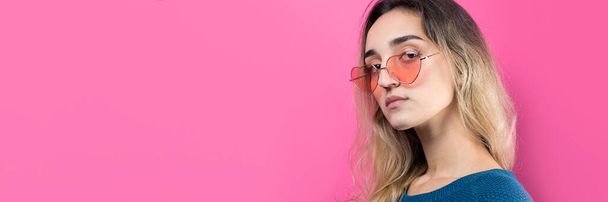 Romantic blonde-haired girl in trendy heart glasses posing with shy smile. Studio close-up portrait on a pink background. - Photo, Image