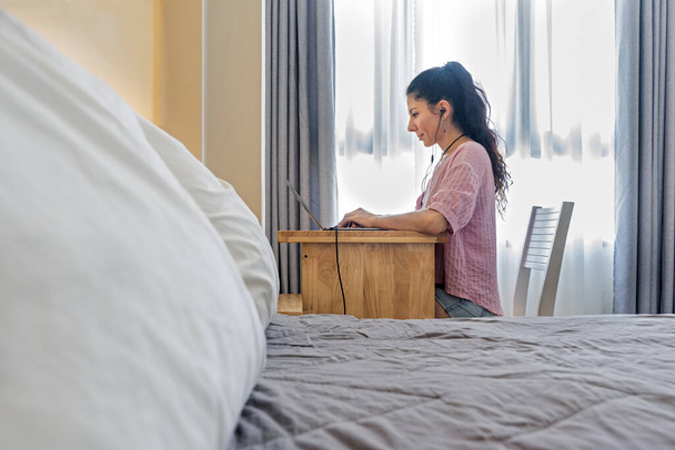 View from behind the bed at a smiling woman working at a laptop in headphones.  Always in touch. Home office concept. Photo with soft selective focus. - Foto, imagen