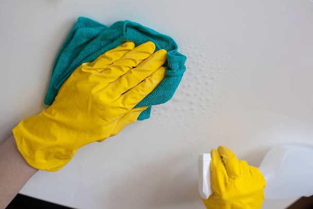 Άνδρας με λαστιχένια γάντια ψεκάζει καθαρότερο σε λευκή επιφάνεια και το σκουπίζει με δαγκάνες. Καθάρισε το σπίτι σου. - Φωτογραφία, εικόνα