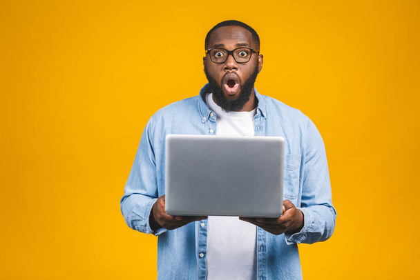 Young surprised african man standing and using laptop computer isolated over yellow background. - Valokuva, kuva