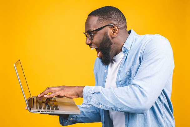 Молодой удивленный африканский американец, стоящий и использующий ноутбук на желтом фоне. - Фото, изображение