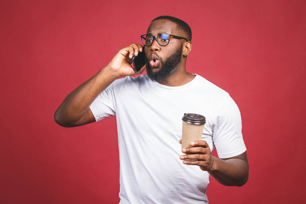 Portré a meglepett jóképű afro-amerikai férfiról, akinek mobiltelefonja van, és elviszi a kávéscsészéjét. Elkülönítve a vörös háttér felett. - Fotó, kép