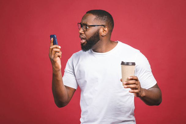 Portré a meglepett jóképű afro-amerikai férfiról, akinek mobiltelefonja van, és elviszi a kávéscsészéjét. Elkülönítve a vörös háttér felett. - Fotó, kép