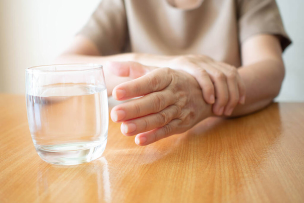 Yaşlı kadın, ahşap masanın üzerindeki bir bardak suya uzanan titreme semptomlarını gösteriyor. El titremesi Parkinson hastalığı, felç ya da beyin hasarı olabilir. Zihinsel sağlık nörolojik bozukluğu. - Fotoğraf, Görsel